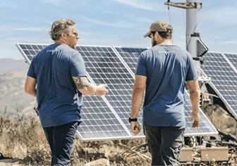 Two people walking along the US/Mexico border near some solar panels.
