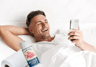 A gleeful man looks at his phone in bed with a bottle of 'Gentleman's Lotion' to his right.